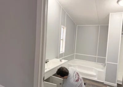 A person working under a white bathroom vanity in a room featuring gray paneled walls and a white bathtub